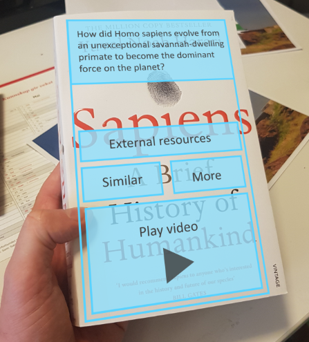 Hand holds a book on which the following AR images are superimposed: 'External resources' button; 'similar' button; 'more' button; 'play video' button.